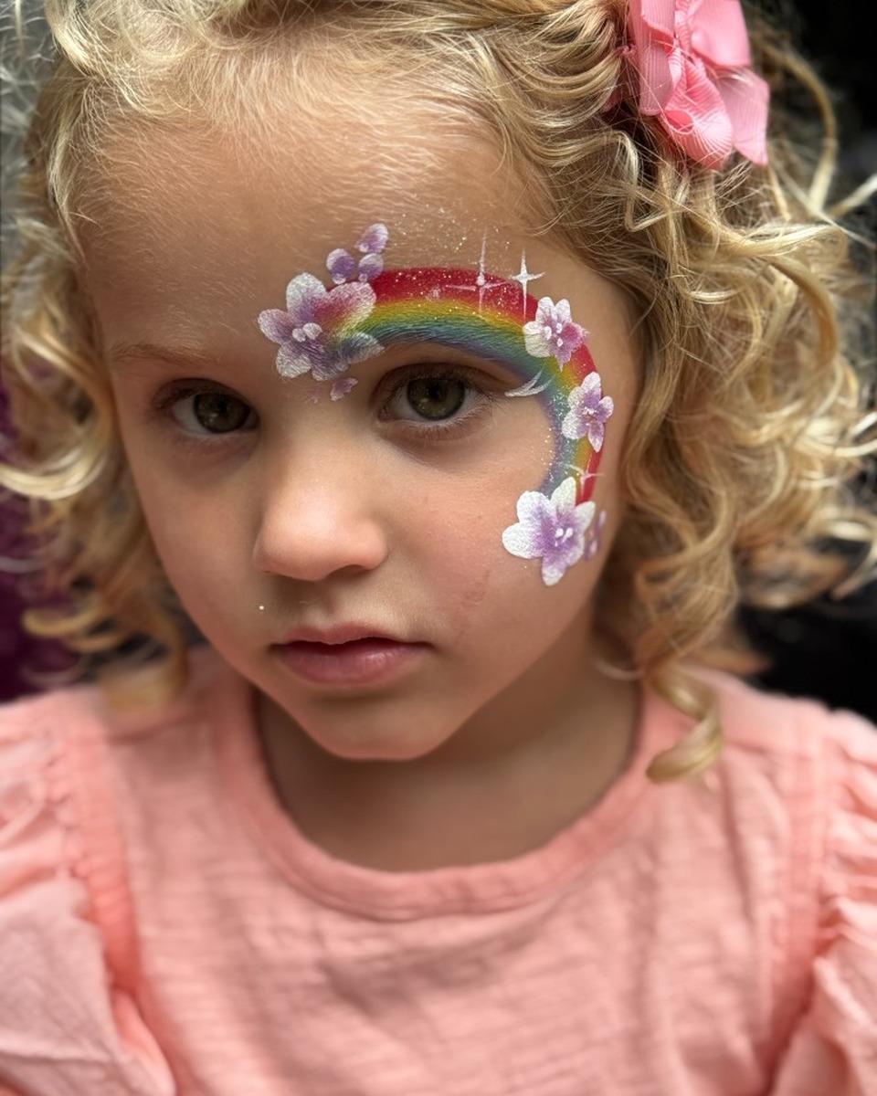 Child with rainbow and flower cheek face painting by Wildflower Face Art